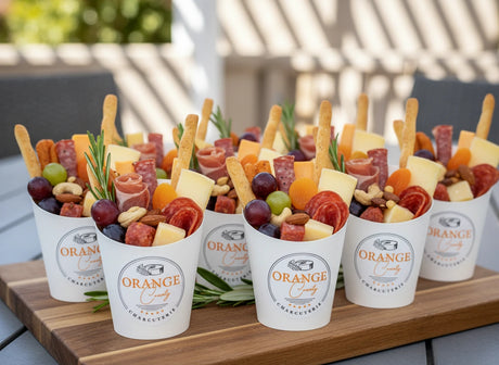 Assorted finger foods in white cups labeled 'Orange County' on a wooden tray.