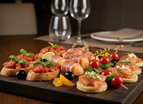 Platter of appetizers with tomatoes, olives, and prosciutto on a wooden board with wine glasses in the background.