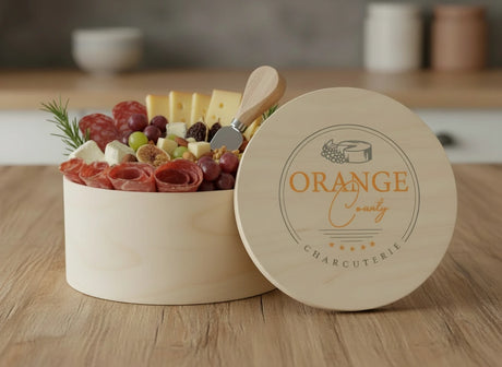 Round charcuterie board with meats, cheeses, and fruits in a wooden box labeled 'Orange County Charcuterie' on a wooden surface.